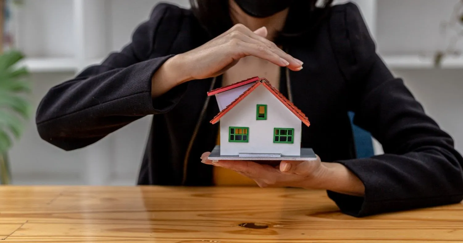 Hands forming a roof over a small model house on a wooden table
